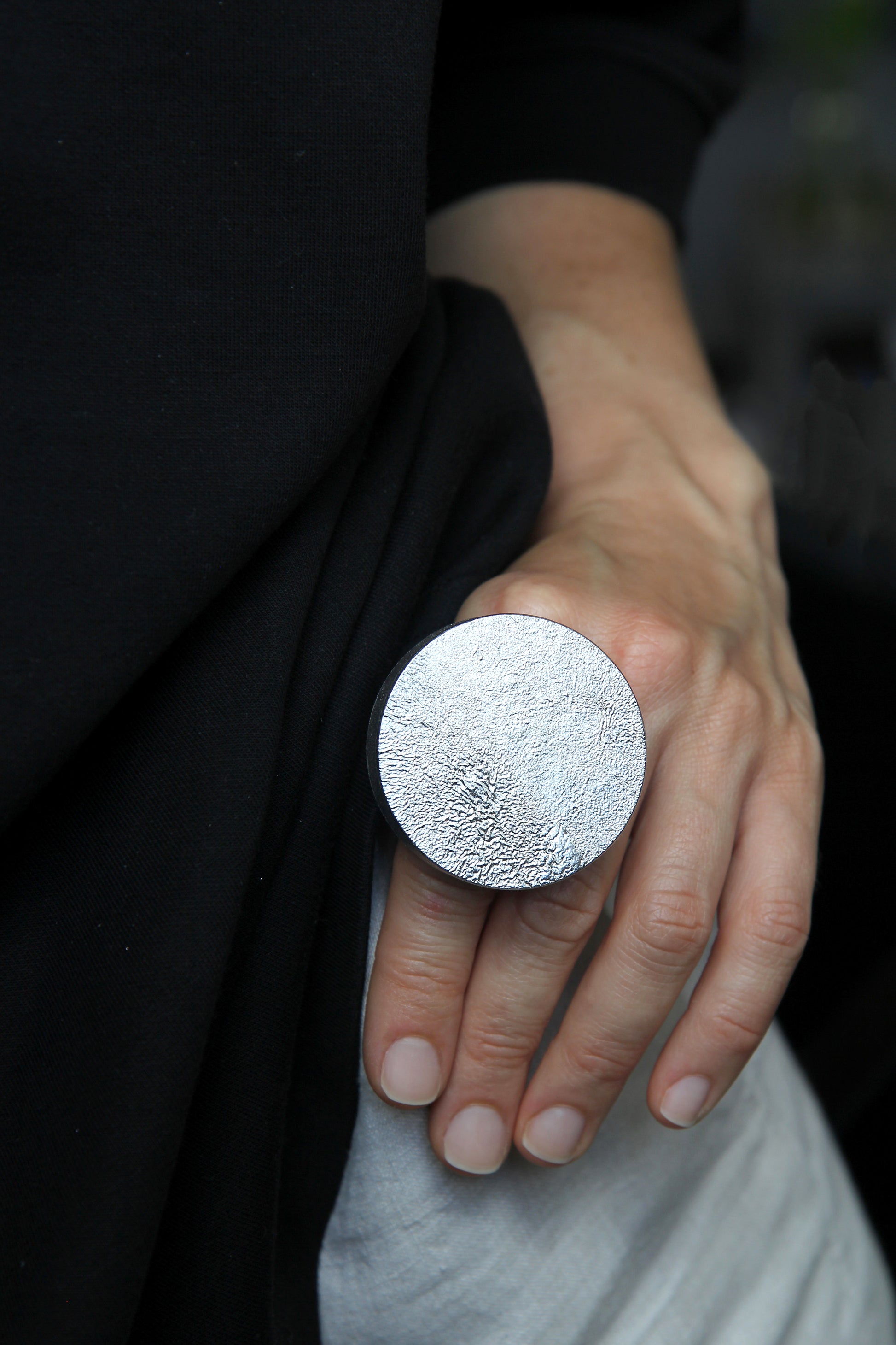 A close-up of a person's hand wearing a large, round, textured silver ring against a dark black top and light gray pants.